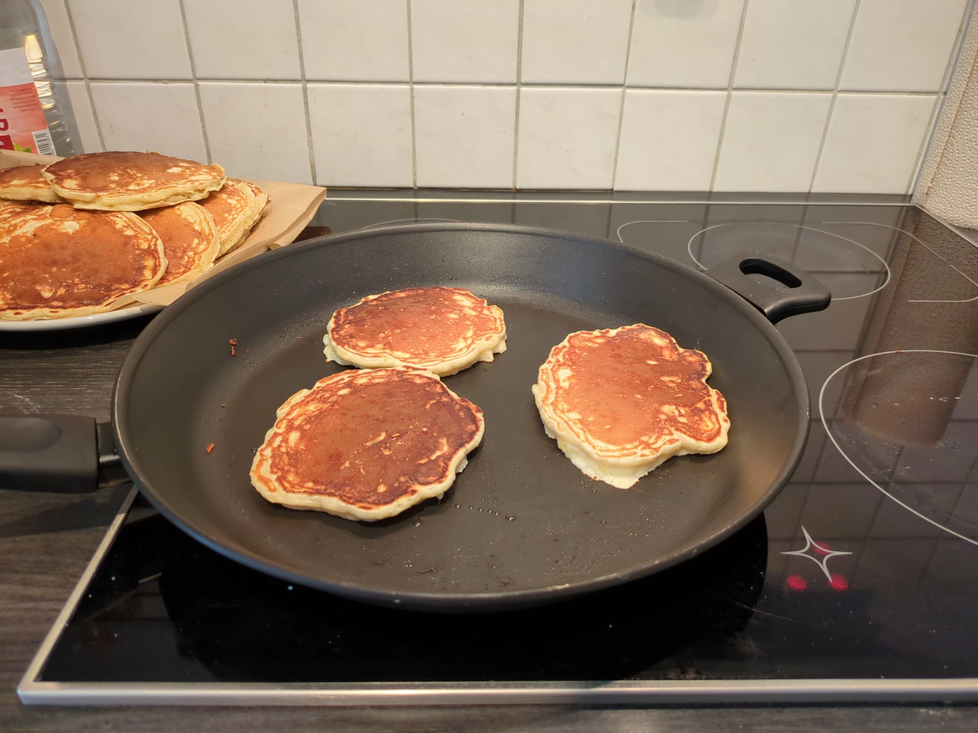 Three pancakes in a large non stick pan on the stove top. A pile of pancakes on a plate off to the side.