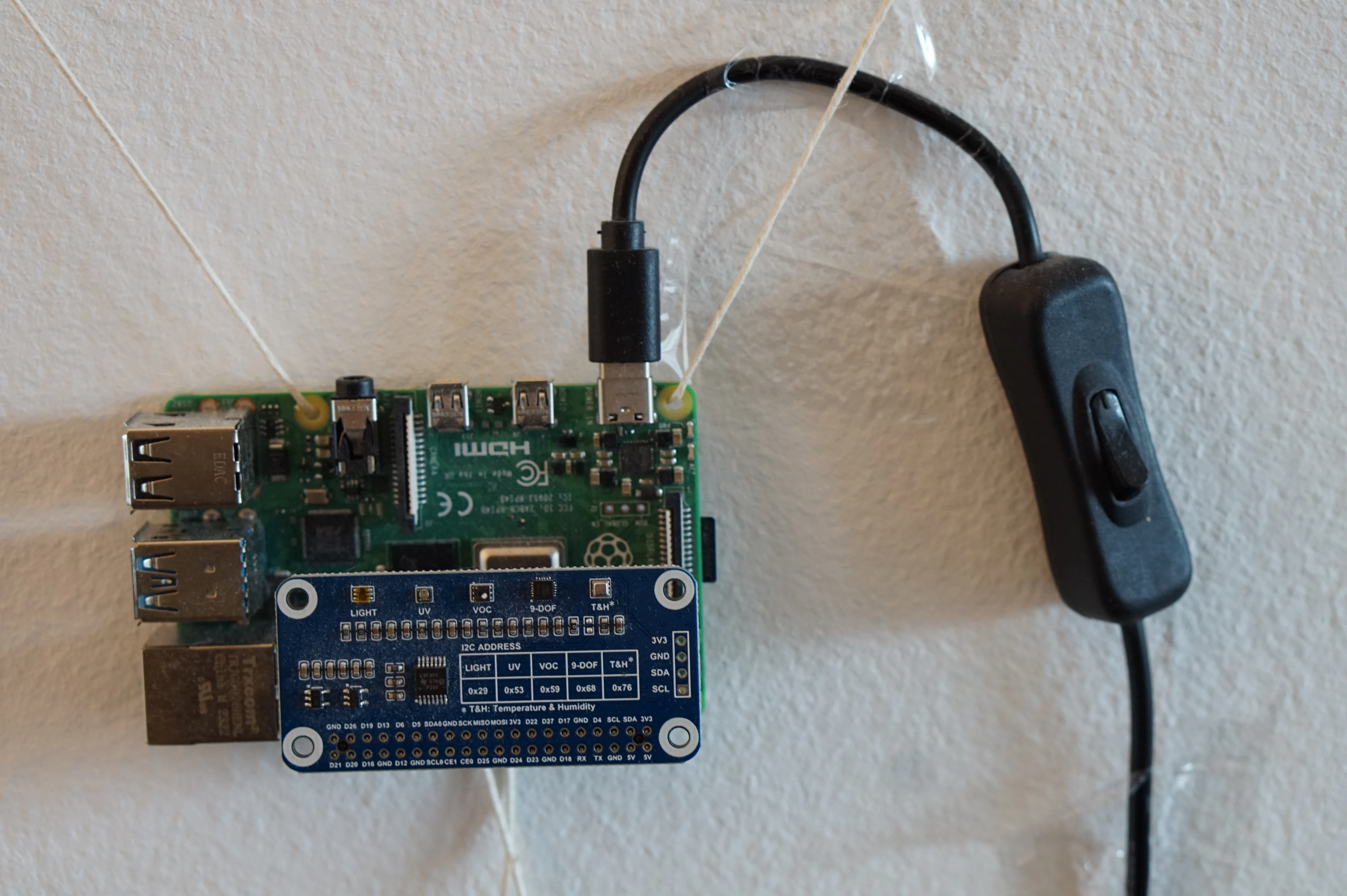 A Raspberry Pi hung on the wall with twine, on two plastic sticky hangers, with a power cord taped to the wall